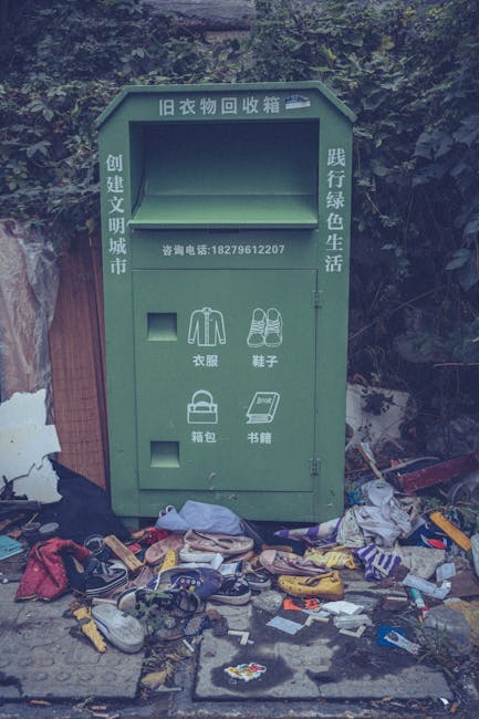 A green metal clothing donation box set against a background of dense bushes and trees, with a large amount of scattered rubbish and discarded items on the ground in front of it. The box features Chin